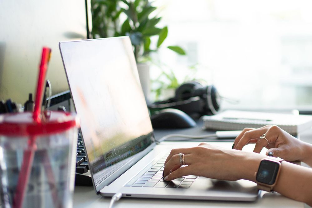 Lady hands type on a laptop. There is a mobile phone on the desk and a large glass of water, clearly staying hydrated.