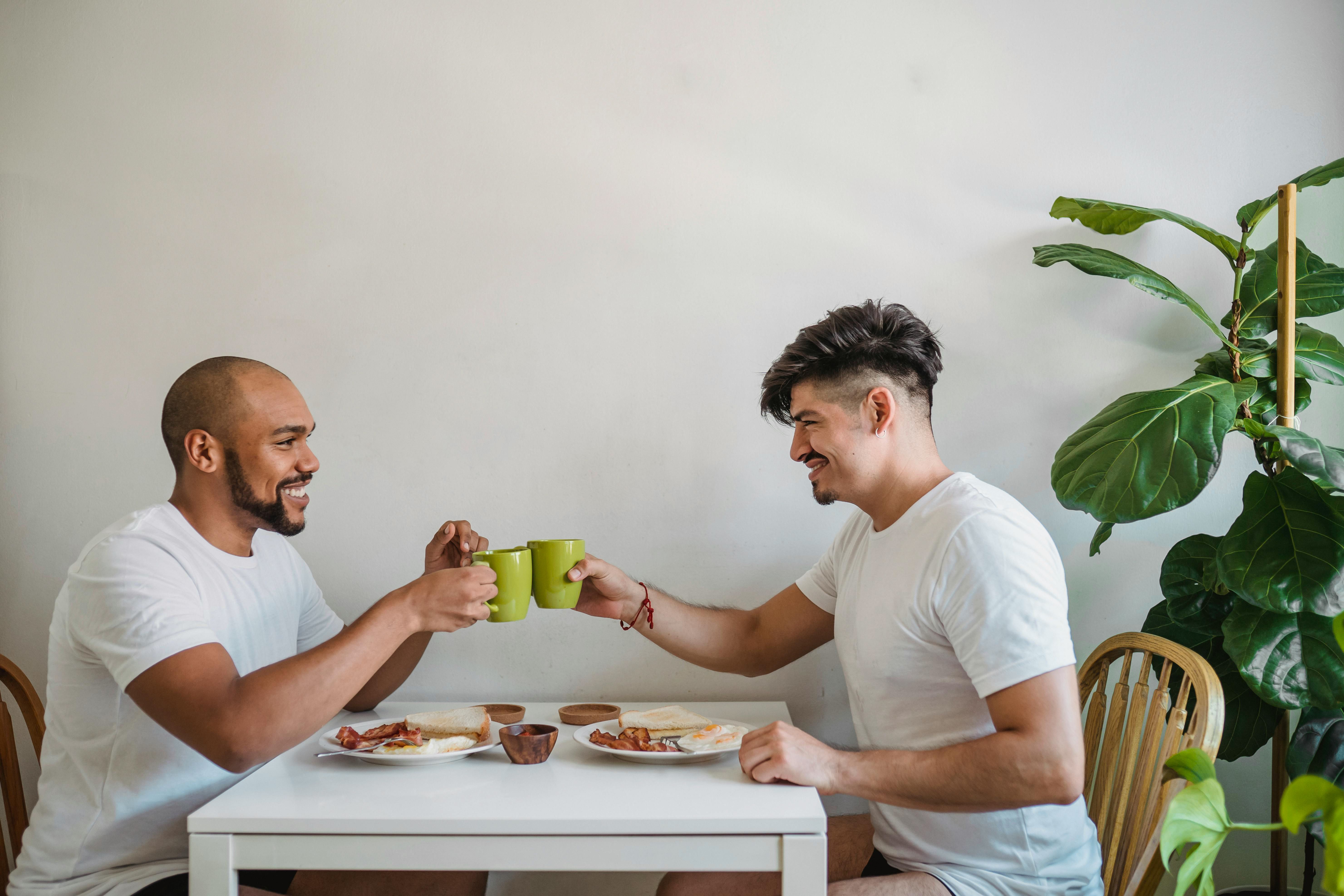 Two men seated at a table over breakfast, toasting with their coffee mugs.