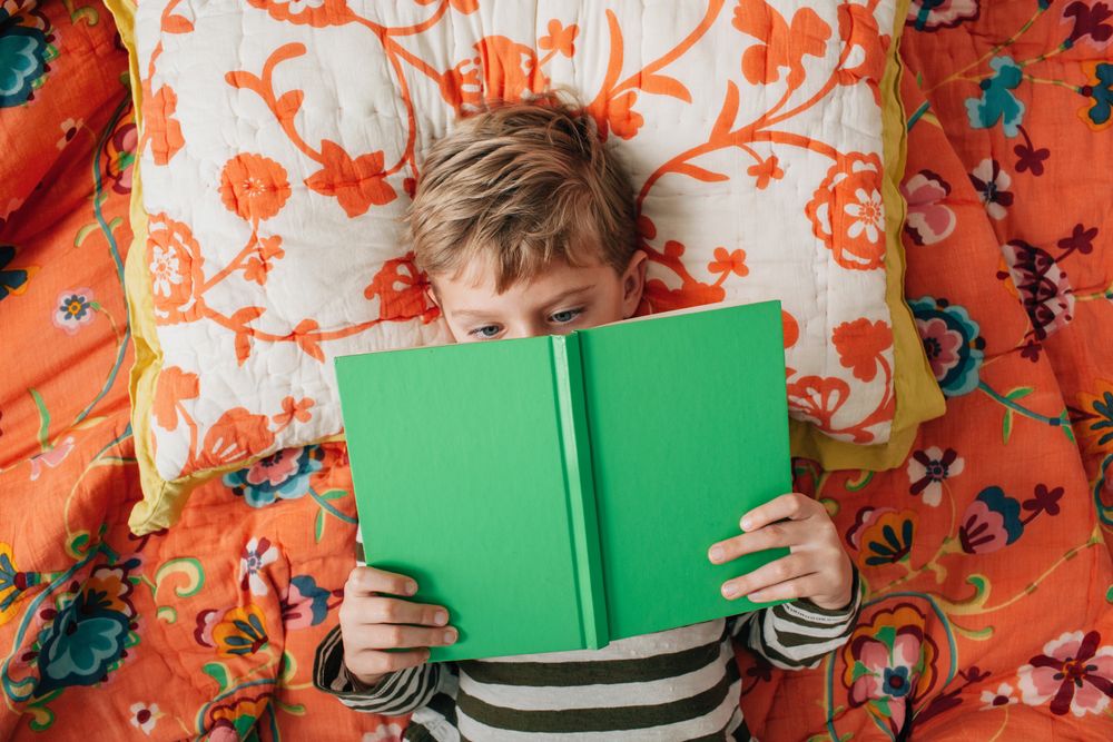 Young blonde boy lying on bed and reading a green book