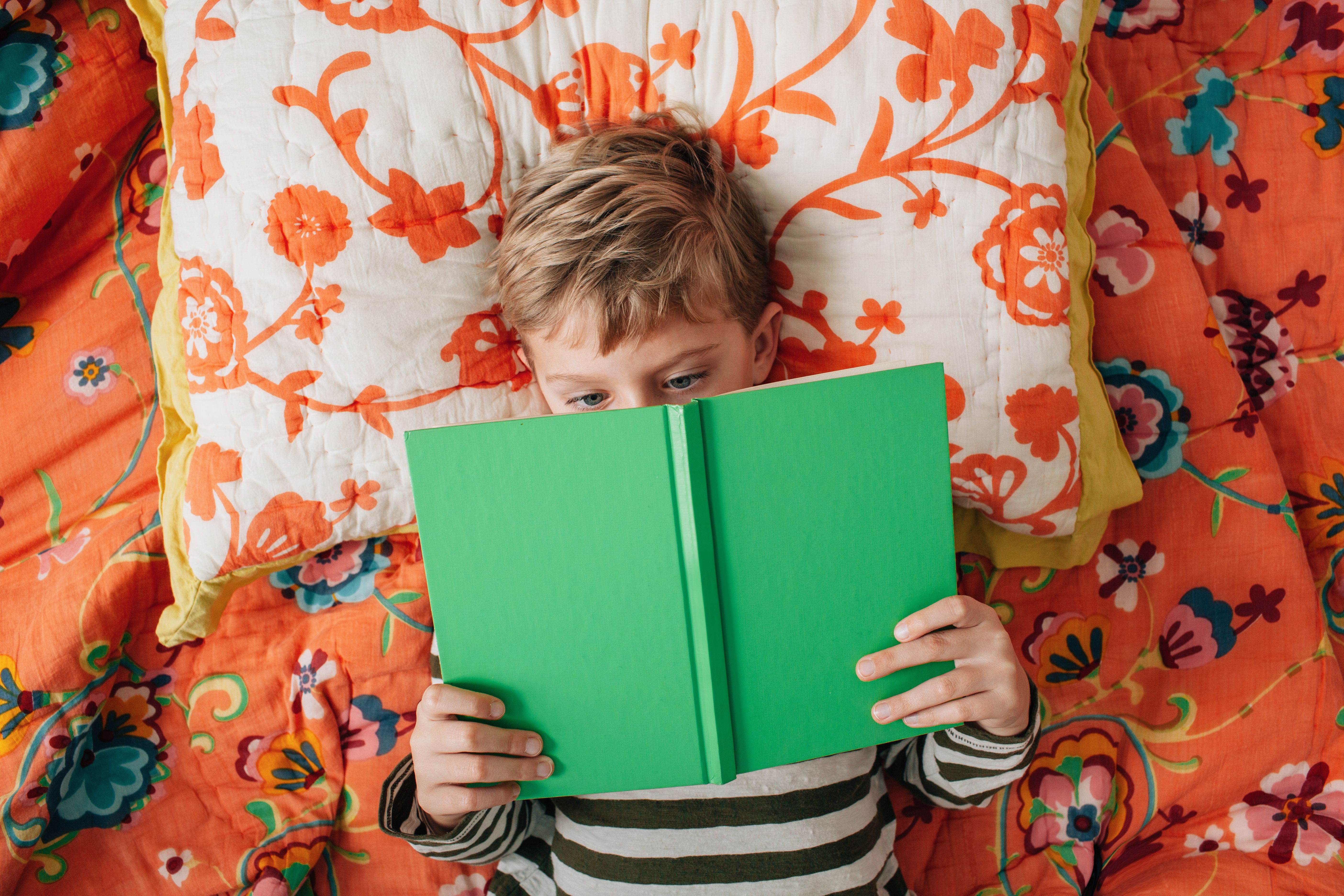 Young blonde boy lying on bed and reading a green book