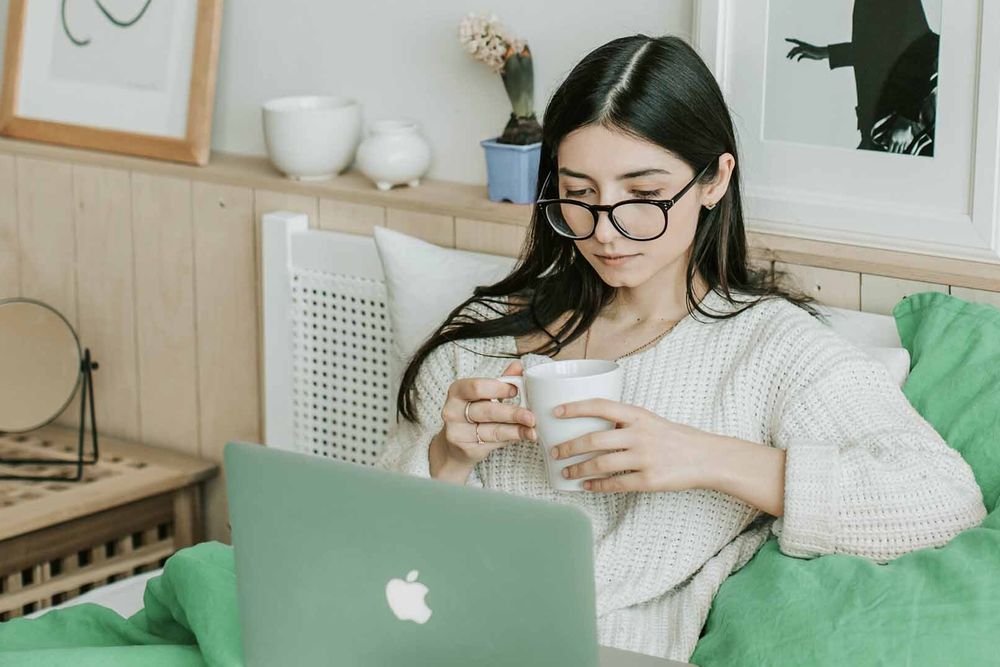 Person sits on their bed reviewing something on their laptop, whilst holding a mug.