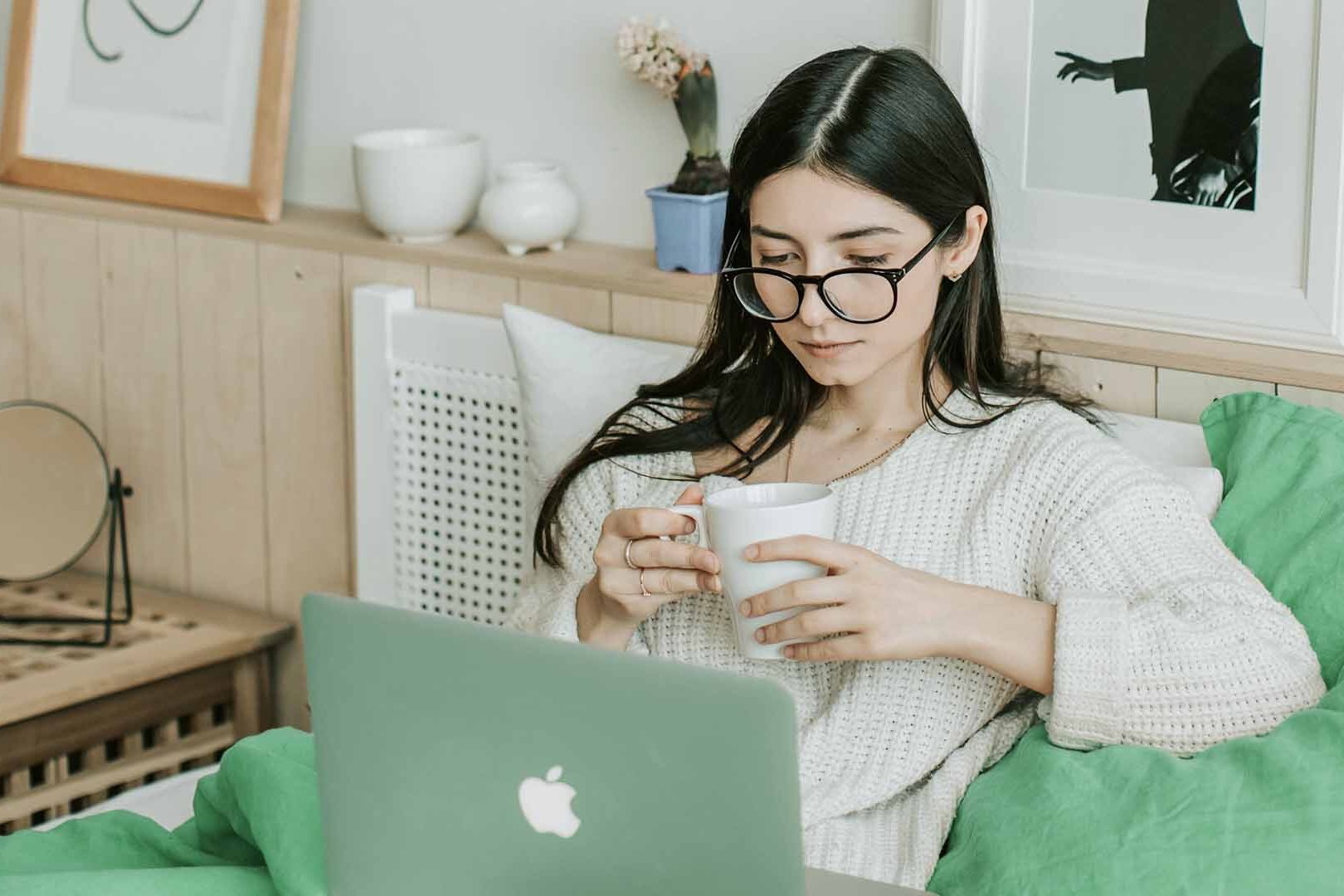 Person sits on their bed reviewing something on their laptop, whilst holding a mug.