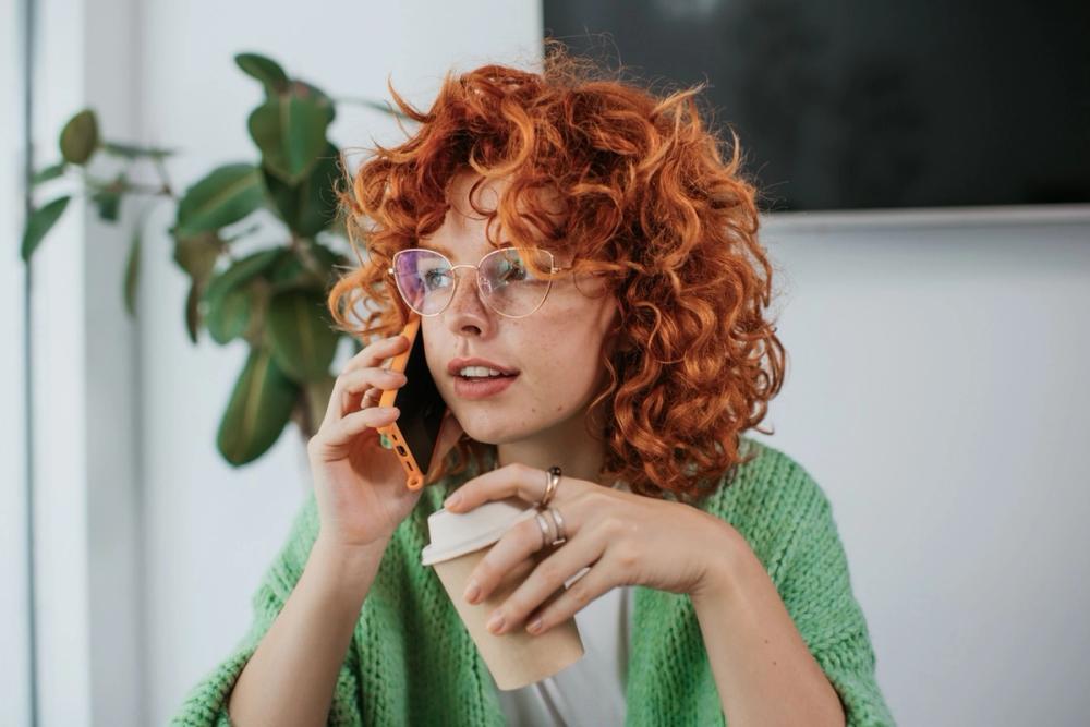 Girl with curly red hair and glasses holding a phone to her ear in one hand and a coffee in the other hand