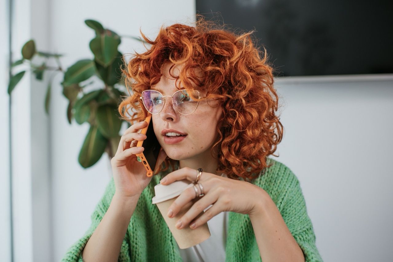 Girl with curly red hair and glasses holding a phone to her ear in one hand and a coffee in the other hand