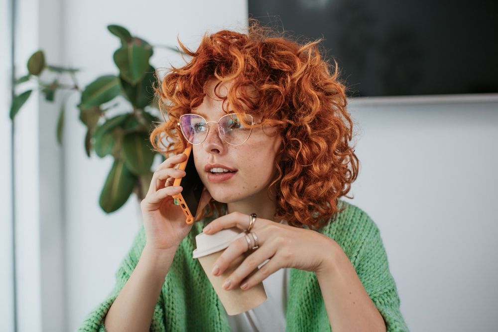 Girl with curly red hair and glasses holding a phone to her ear in one hand and a coffee in the other hand