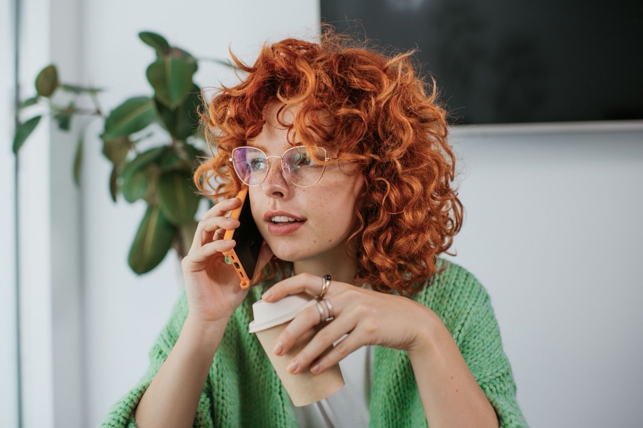 Girl with curly red hair and glasses holding a phone to her ear in one hand and a coffee in the other hand