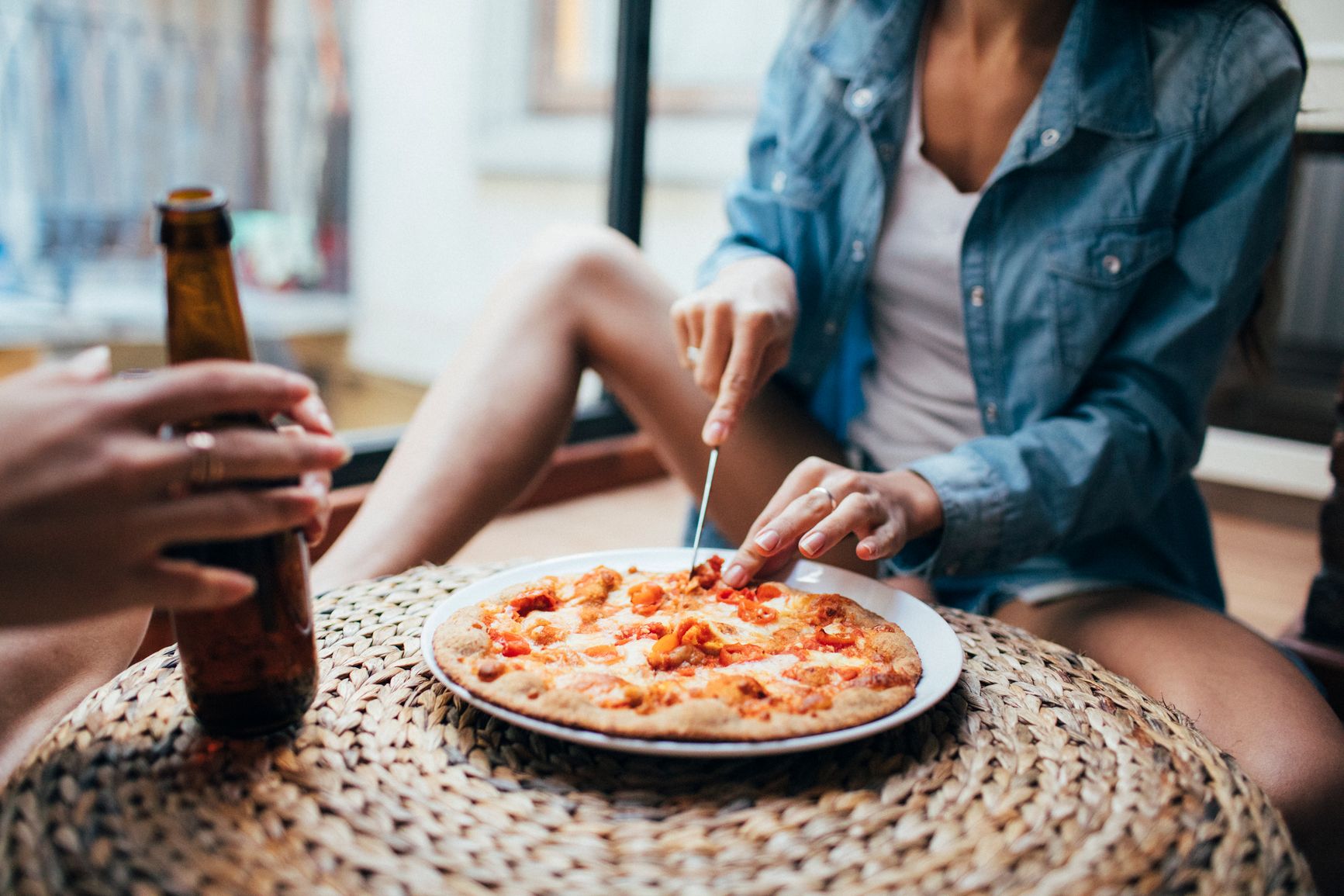 A woman seated on the floor in a home, cutting a whole pizza on the ottoman.