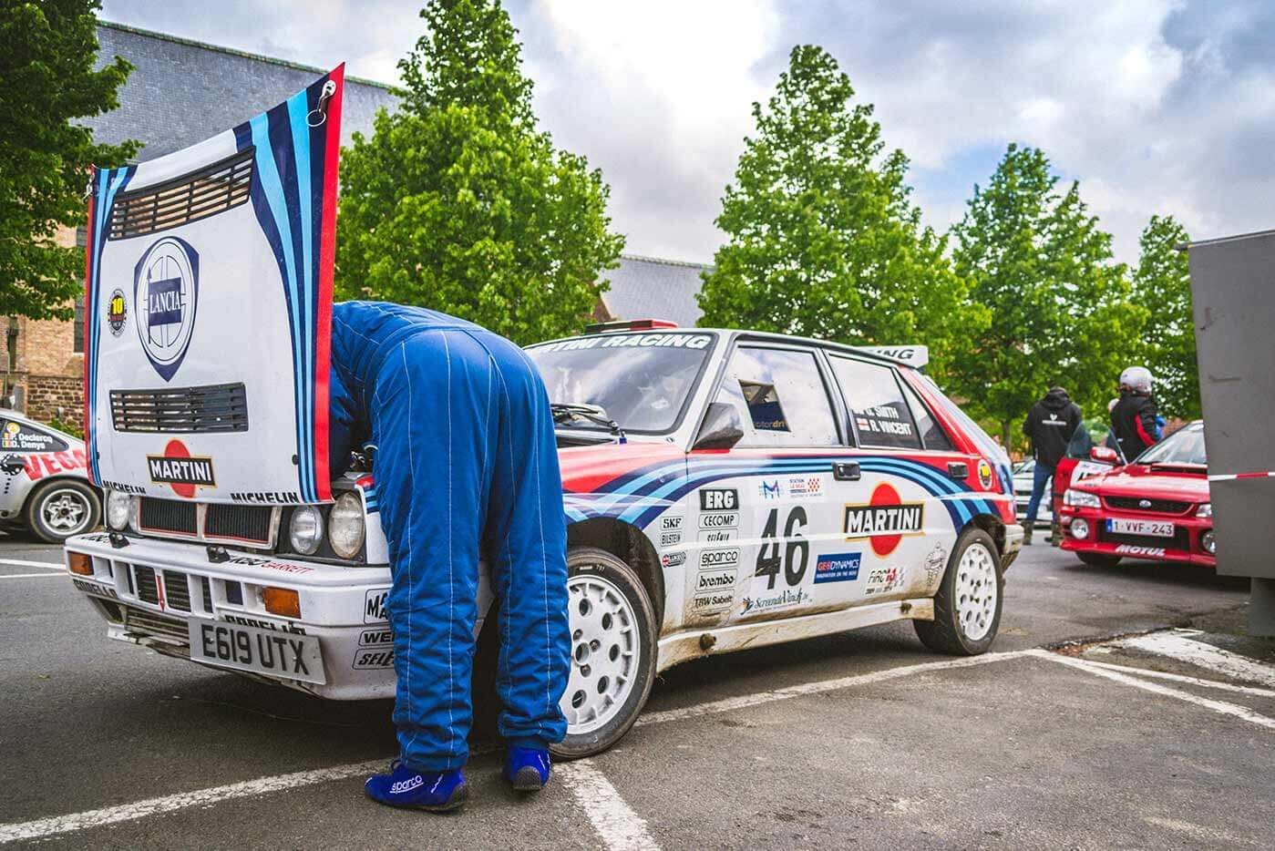 An Audi dirt racing car sits in a carpark with its hood up. A mechanic is shoulders deep in the engine bay looking at something unknown.