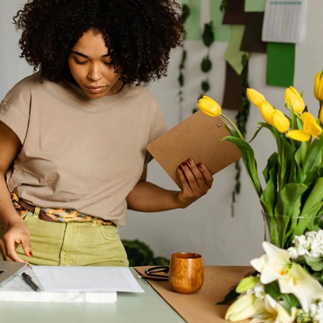 A woman looks at a piece of paper, whilst holder a small wooden clipboard. There are tulips in a vase on the desk.