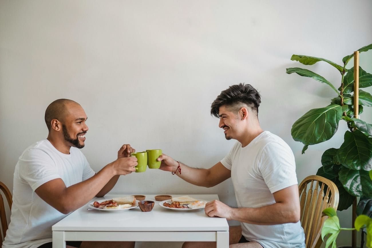 Two men seated at a table over breakfast, toasting with their coffee mugs.