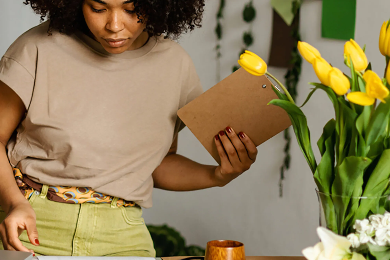 A woman looks at a piece of paper, whilst holder a small wooden clipboard. There are tulips in a vase on the desk.