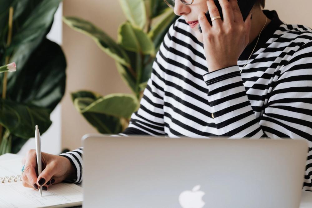 Woman sitting in front of her laptop on her mobile and taking notes.