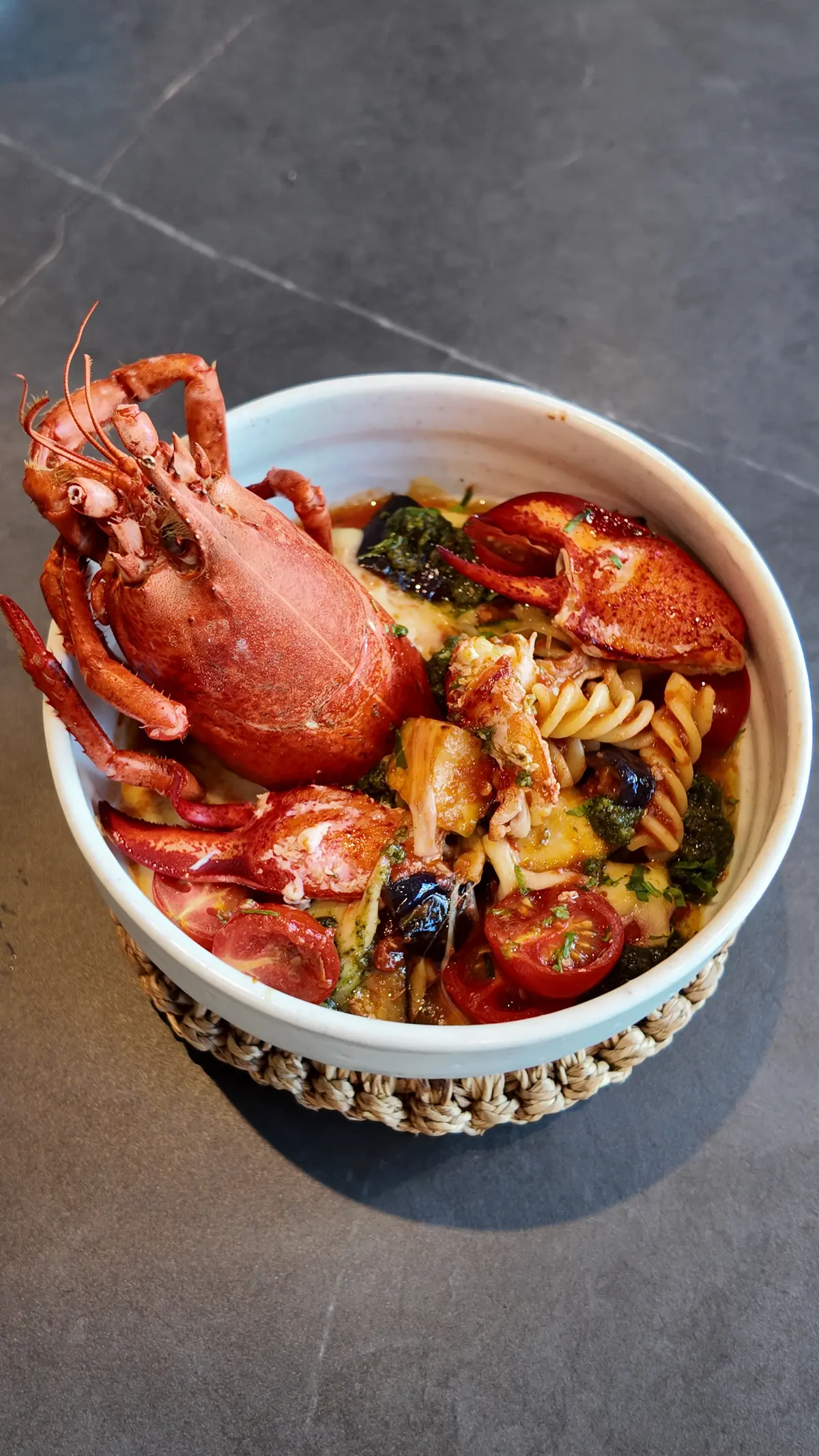 Whole lobster served over fusilli pasta with cherry tomatoes, pesto and olives in a ceramic bowl on stone