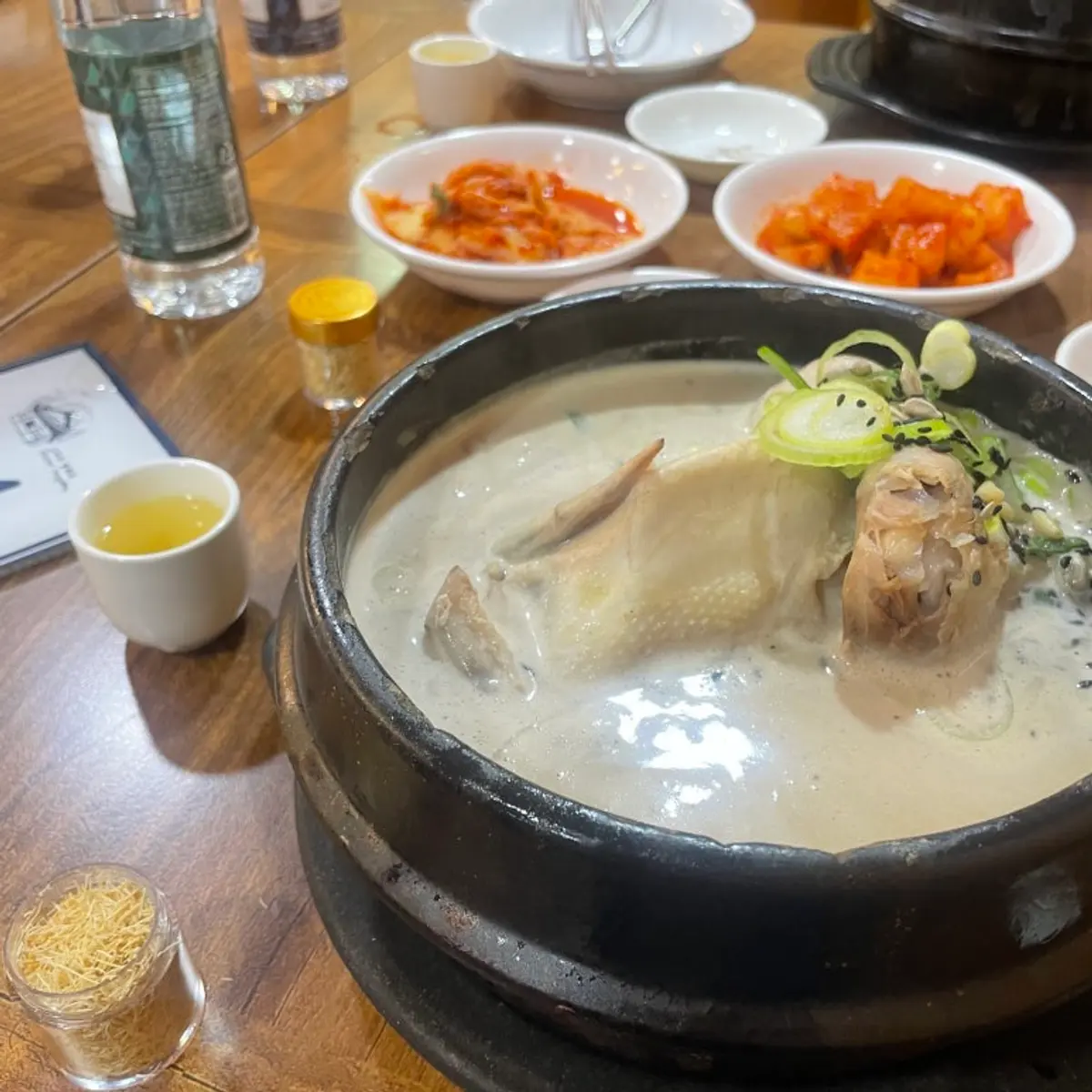 김치 반찬과 인삼주가 곁들여진, 검은 돌솥에 담긴 삼계탕
