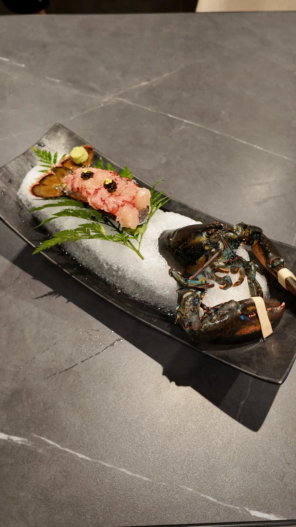 Live lobster sashimi presentation with caviar, wasabi, and fern on crushed ice over a black ceramic plate