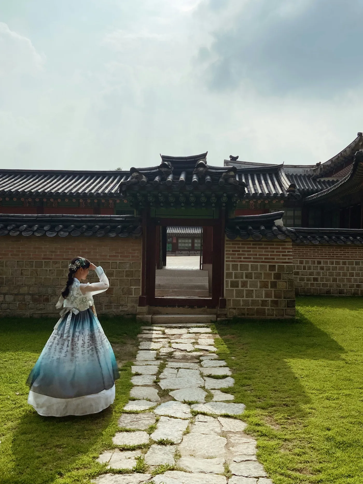푸른 그라데이션 한복을 입은 소녀가 서울의 기와 궁궐 문으로 이어지는 돌길에서 잠시 멈춰 서 있다