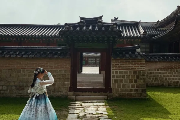 A girl in a blue gradient hanbok pauses by a stone path leading to a tiled palace gate in Seoul