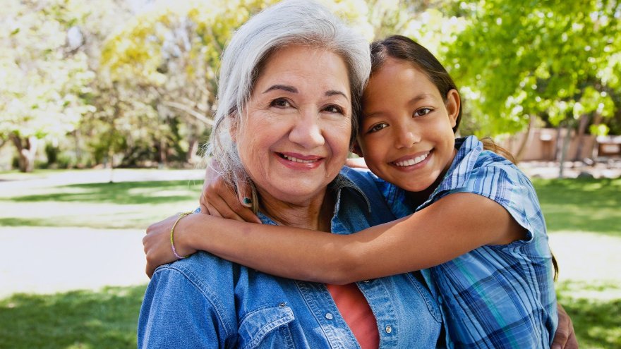 A grandmother and her grandchild smile at the camera outdoors while hugging.