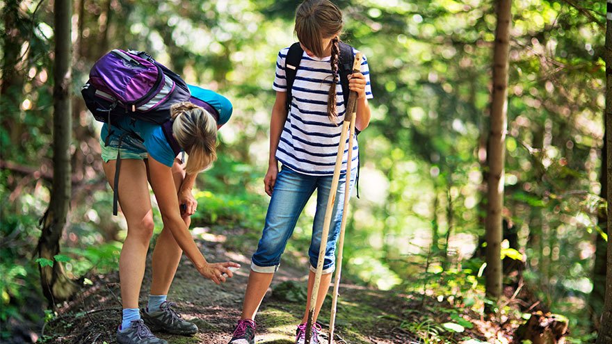 Mom and daughter hiking in forest as mom applies tick repellent on the daughter's legs