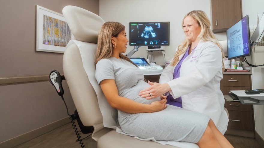 A pregnant woman sits in an exam chair while her OB/GYN touches her stomach.