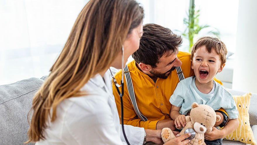 Doctor examining small boy with stethoscope in the office, with his father sitting next to him.