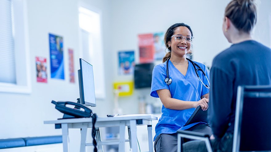 A provider sits at her desk with a female patient across from her as they discuss her concerns