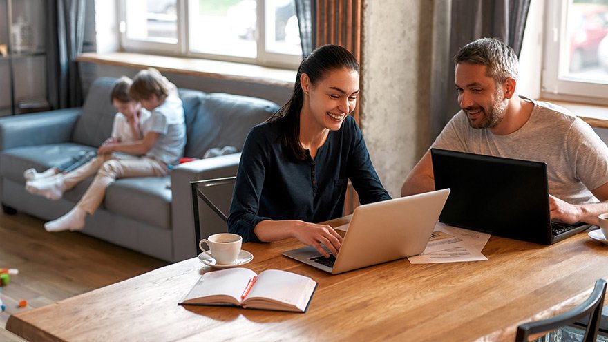 Couple doing paperwork on laptops while children play in background