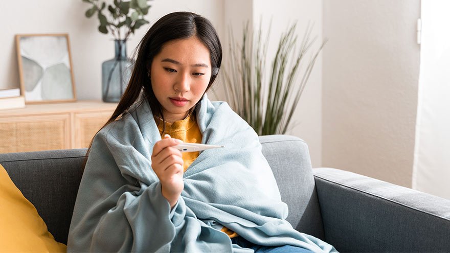 Sick woman at home covered with a blanket while using thermometer to take her temperature.