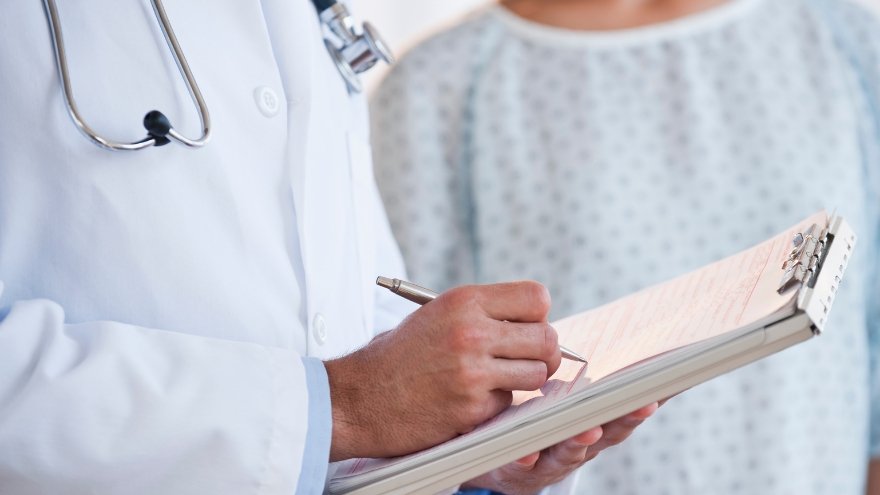 A doctor writes notes on a clipboard next to a patient in a gown.