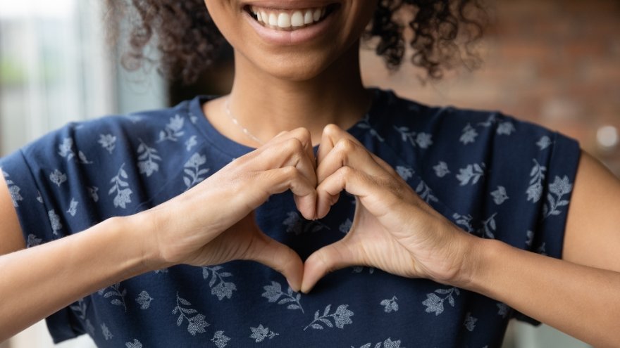 A woman in a blue, floral print shirt puts her hands together to make a heart while smiling for the camera.