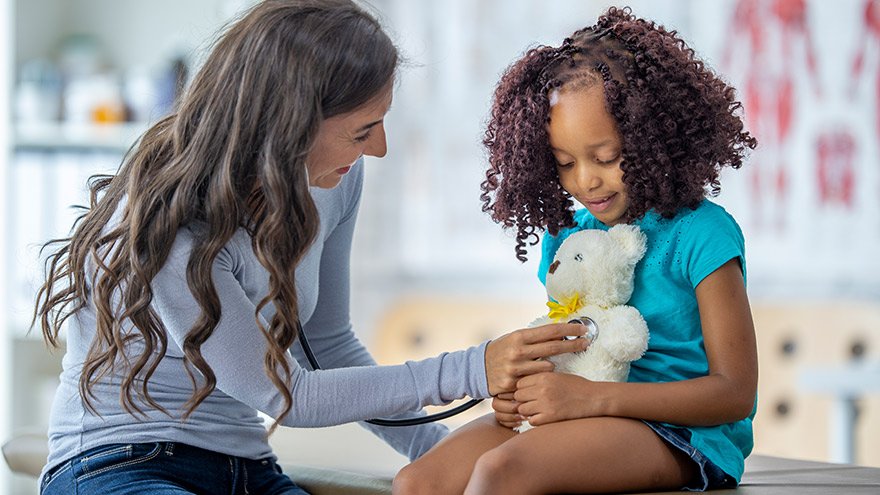 A young girl on a medical exam table as her doctor listens to her teddy bears heart.