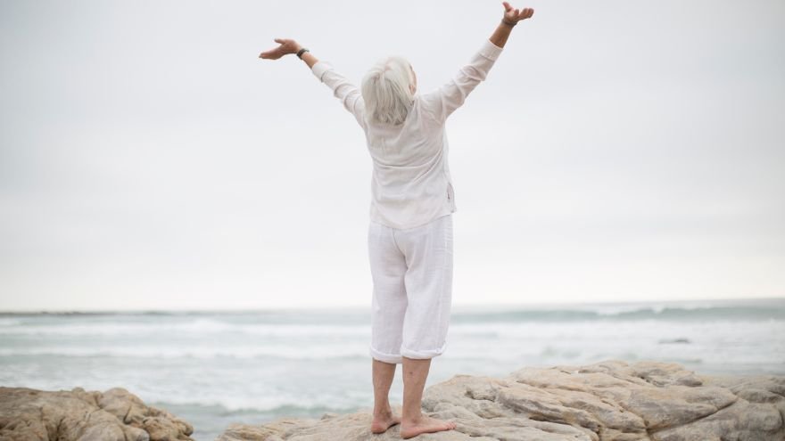 An elderly woman standing on top of rocks at a beach with arms extended