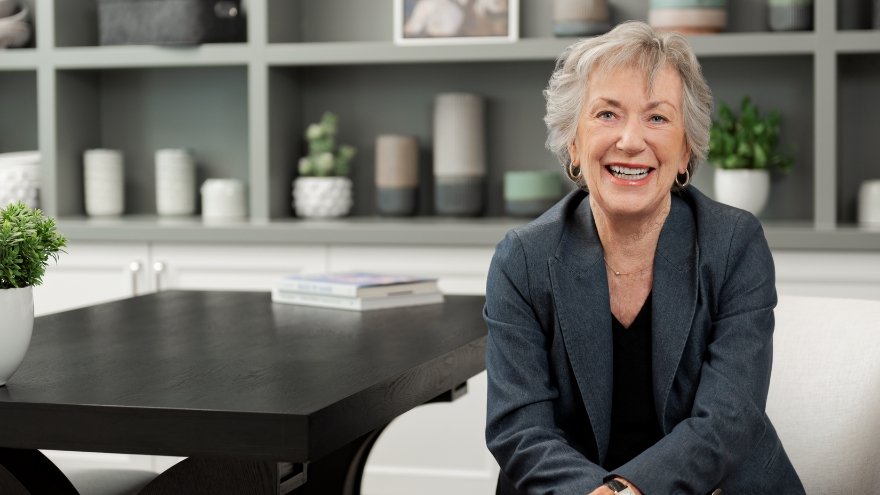 Senior Care Plus member JoAnne Conley sits on a couch inside a home and smiles for the camera.