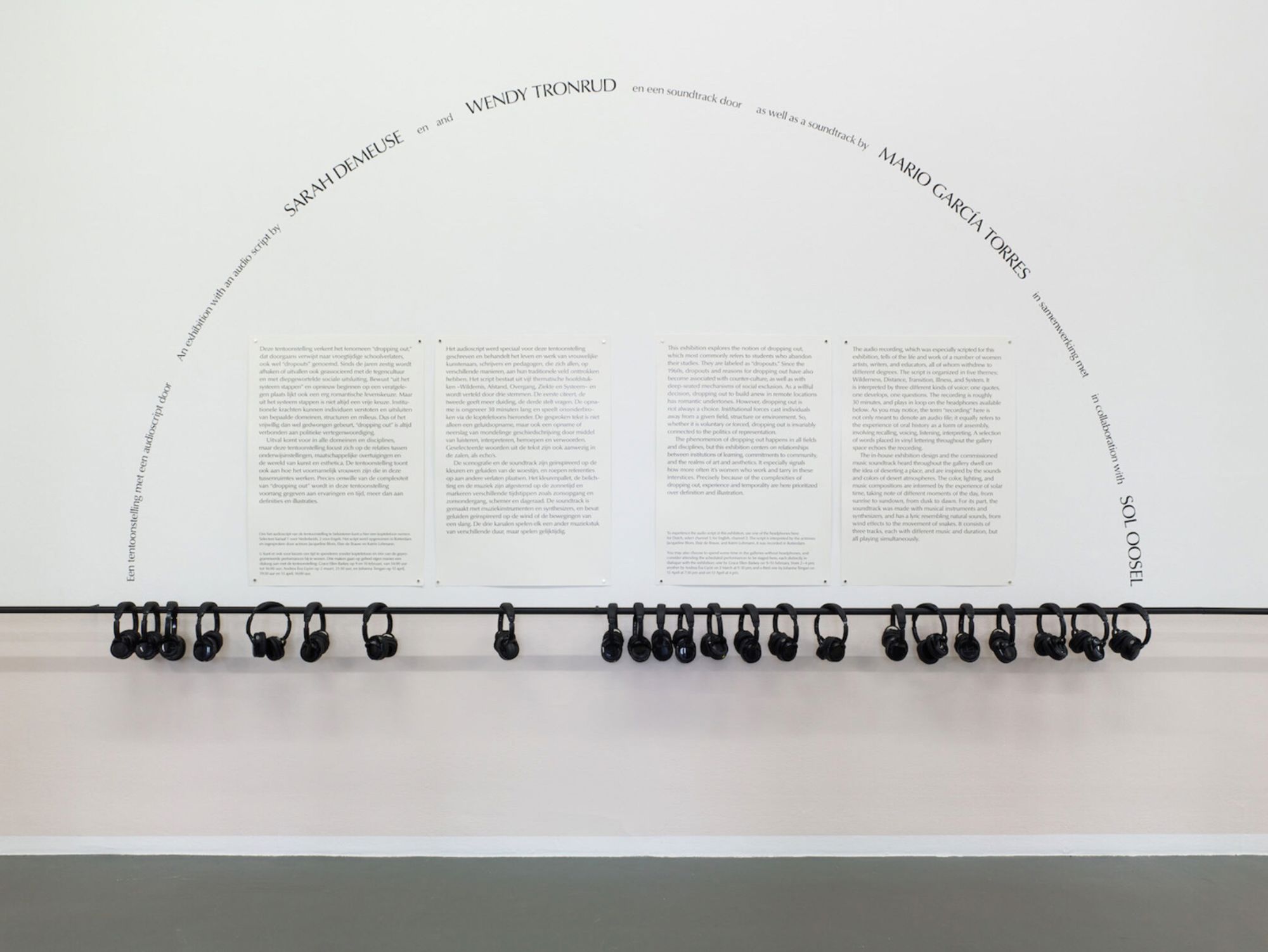 Straight-on photo of a white wall with black exhibition wall text laid out in four columns, with the exhibition title laid out in an arch around it. On a horizontal bar attached to the wall are 25 wireless headphones. The bottom of the wall is painted light pink.