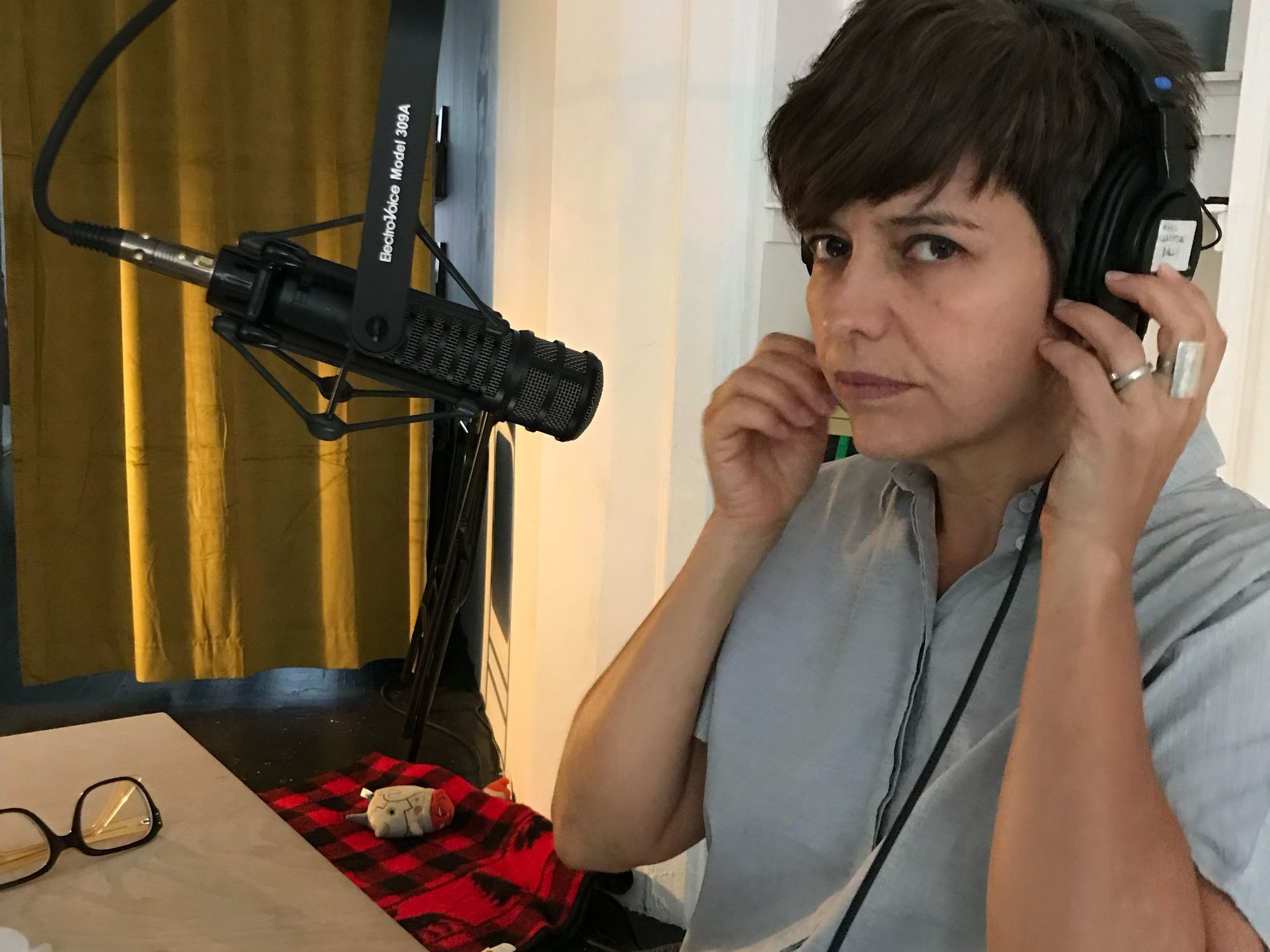 A person with short brown hair and a light blue shirt, putting on headphones and sitting in front of a microphone in a recording studio