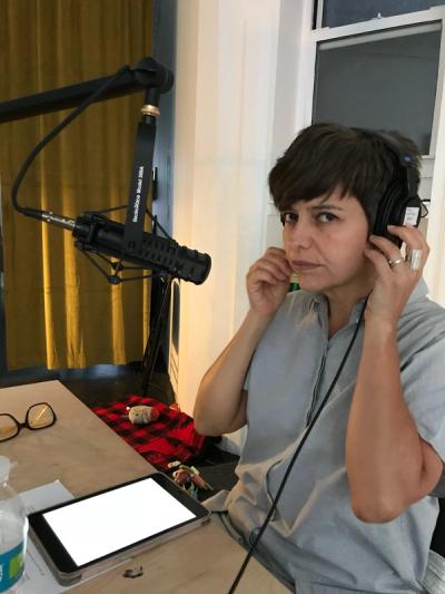 A person with short brown hair and a light blue shirt, putting on headphones and sitting in front of a microphone in a recording studio