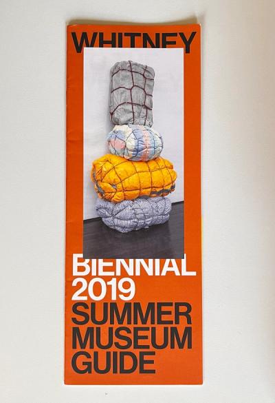 Scan of the front of a summer 2019 visitor guide to the Whitney Biennial, cover is orange with black and white text in caps and a photo of an artwork.