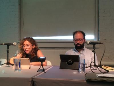 Two people sitting behind a table, they read from laptops and sit in front of microphones.