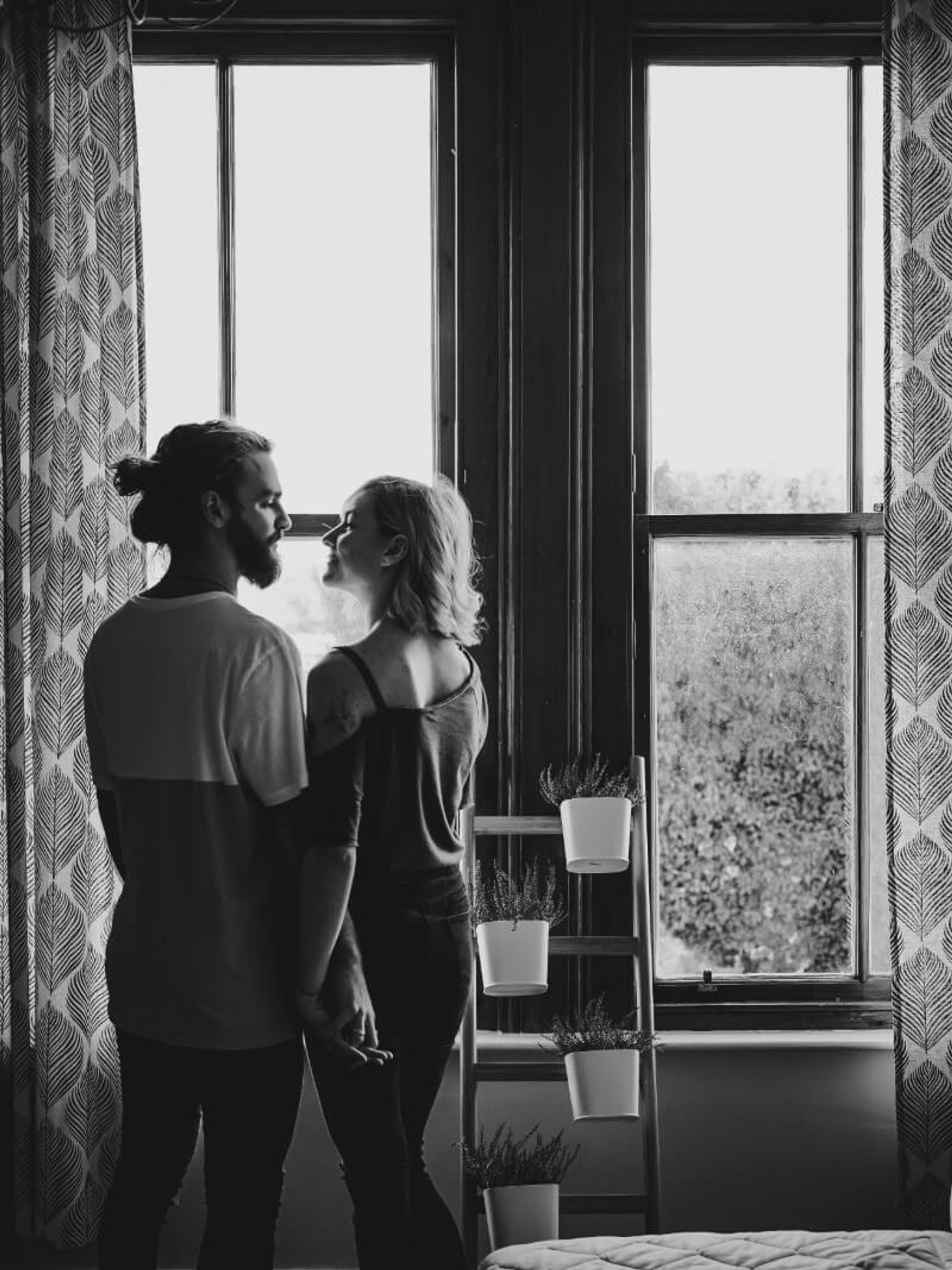 A couple stands together by a window, holding hands and facing each other.