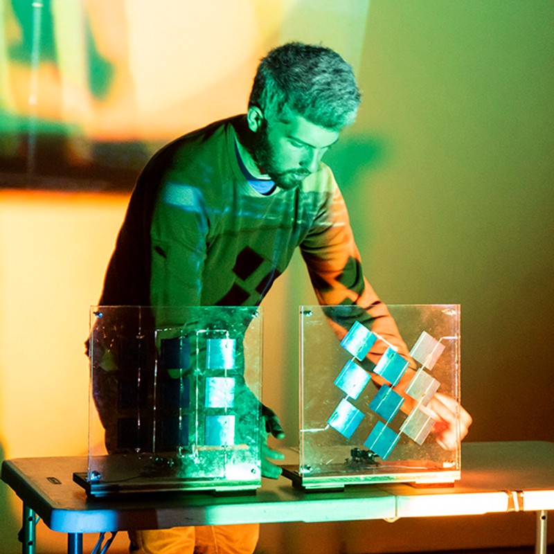 An early-30s man in a long sleeve shirt stands behind a table with two photovoltaic sculptures on it. Green light is being projected on the man and table.