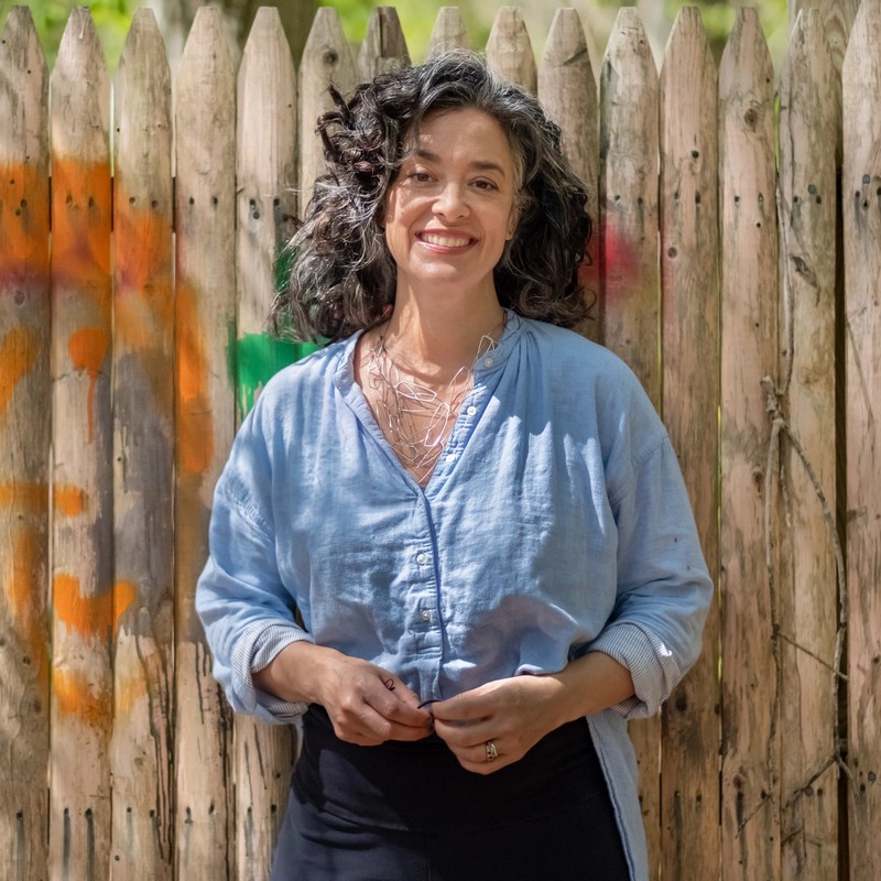 Photo of Kara Gilmour, a white woman with silver and black hair, in a light blue shit with an intersecting silver necklace and a broad smile. She stands in front of a wooden fence with brightly colored children's graffiti adorning it.