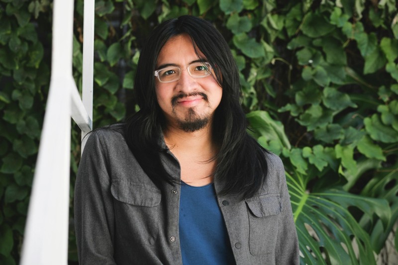 Headshot of Miguel A. López with long black hair and wearing glasses, in front of a green background