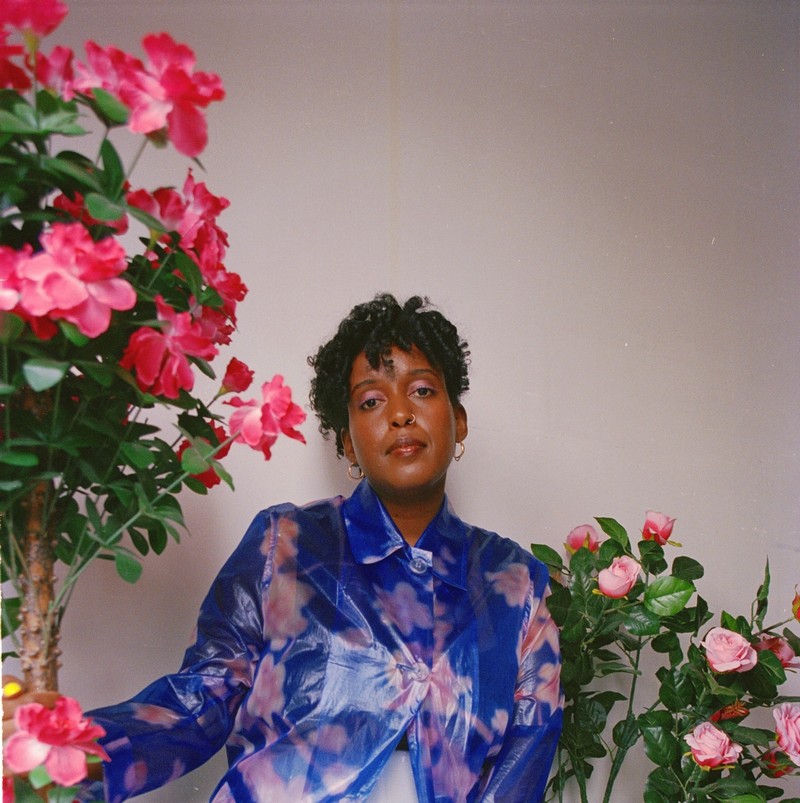 Headshot of Salome Asega with curly bangs and wearing gold hoops, nose ring, and a blue and pink floral jacket, sitting beside bright pink flowers.