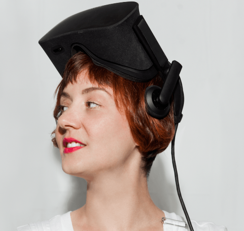 Headshot of Amelia Winger-Bearskin a woman with short brown hair and wearing a virtual reality headset on top of her head while looking sideways, wearing white overalls and red lipstick.