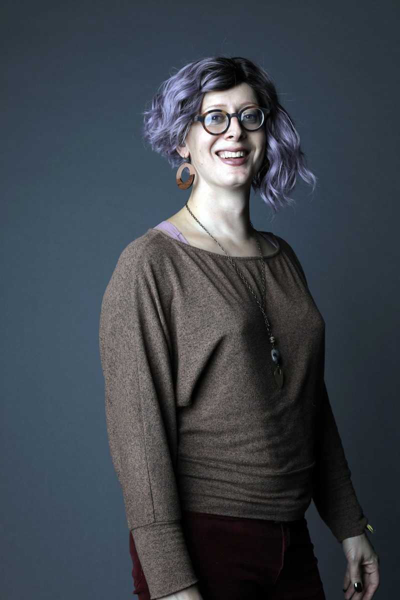 Headshot of Sacha with asymmetrical lavender hair and wearing glasses, in front of a greyish blue background.