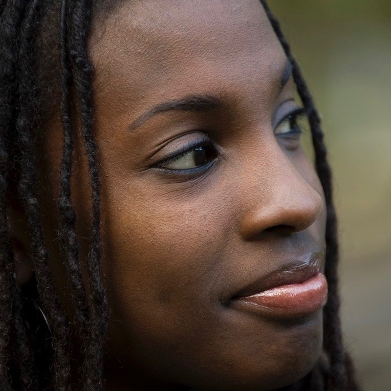A young woman smiles contently, gazing into the distance to her left. She has dark brown skin and thin black locs framing either side of her face.