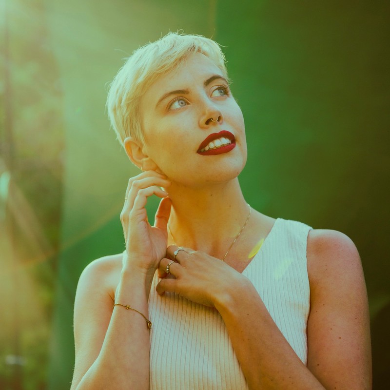 Headshot of Paige with short blonde hair, red lipstick, looking upward, in front of a green backdrop with lens flair.
