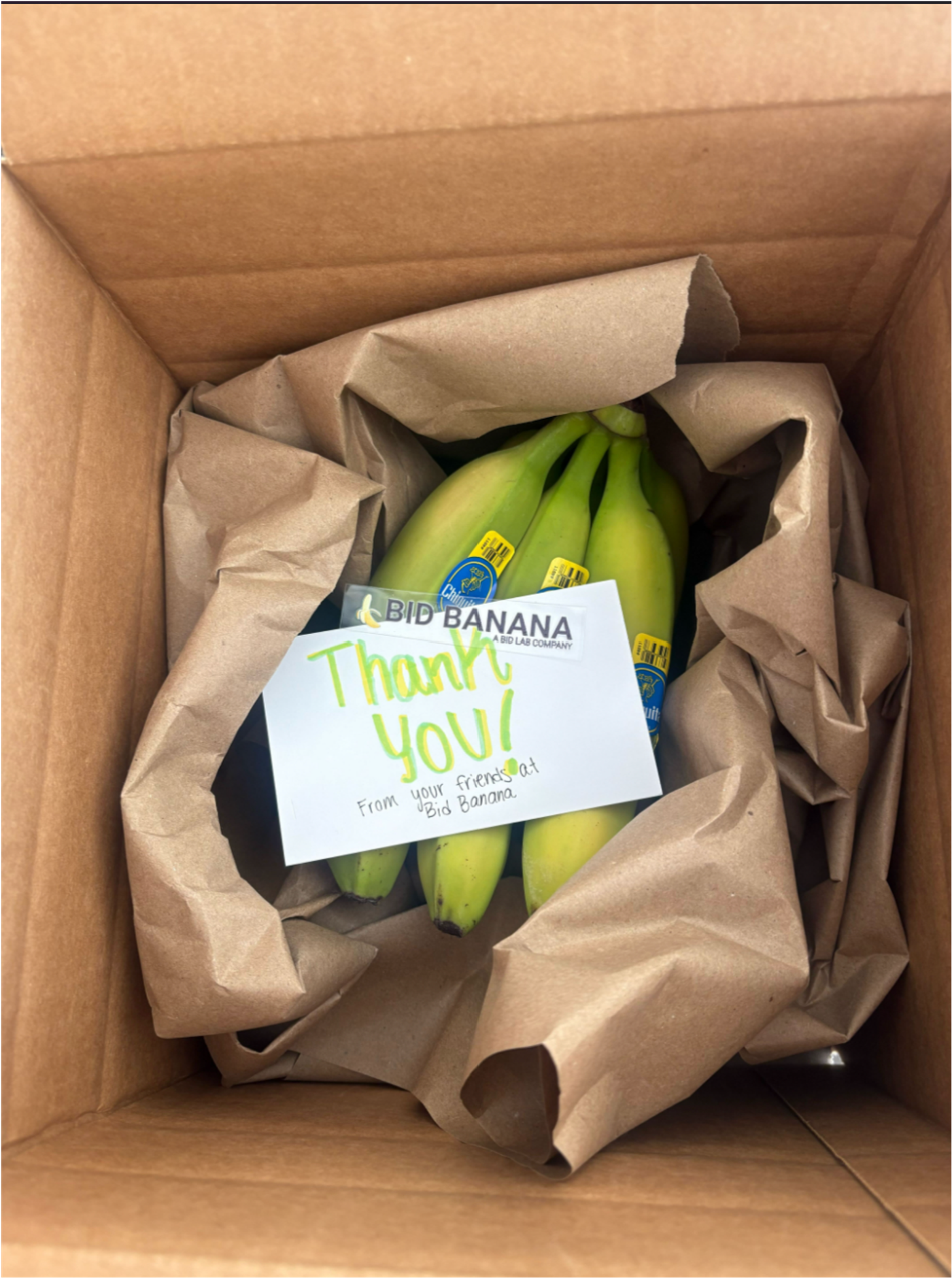 An open cardboard box of green bananas nestled in brown paper, with a handwritten card: “Thank you! From your friends at Bid Banana.”