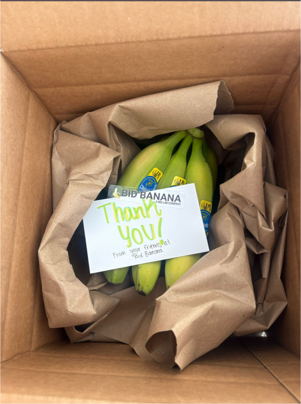 An open cardboard box of green bananas nestled in brown paper, with a handwritten card: “Thank you! From your friends at Bid Banana.”