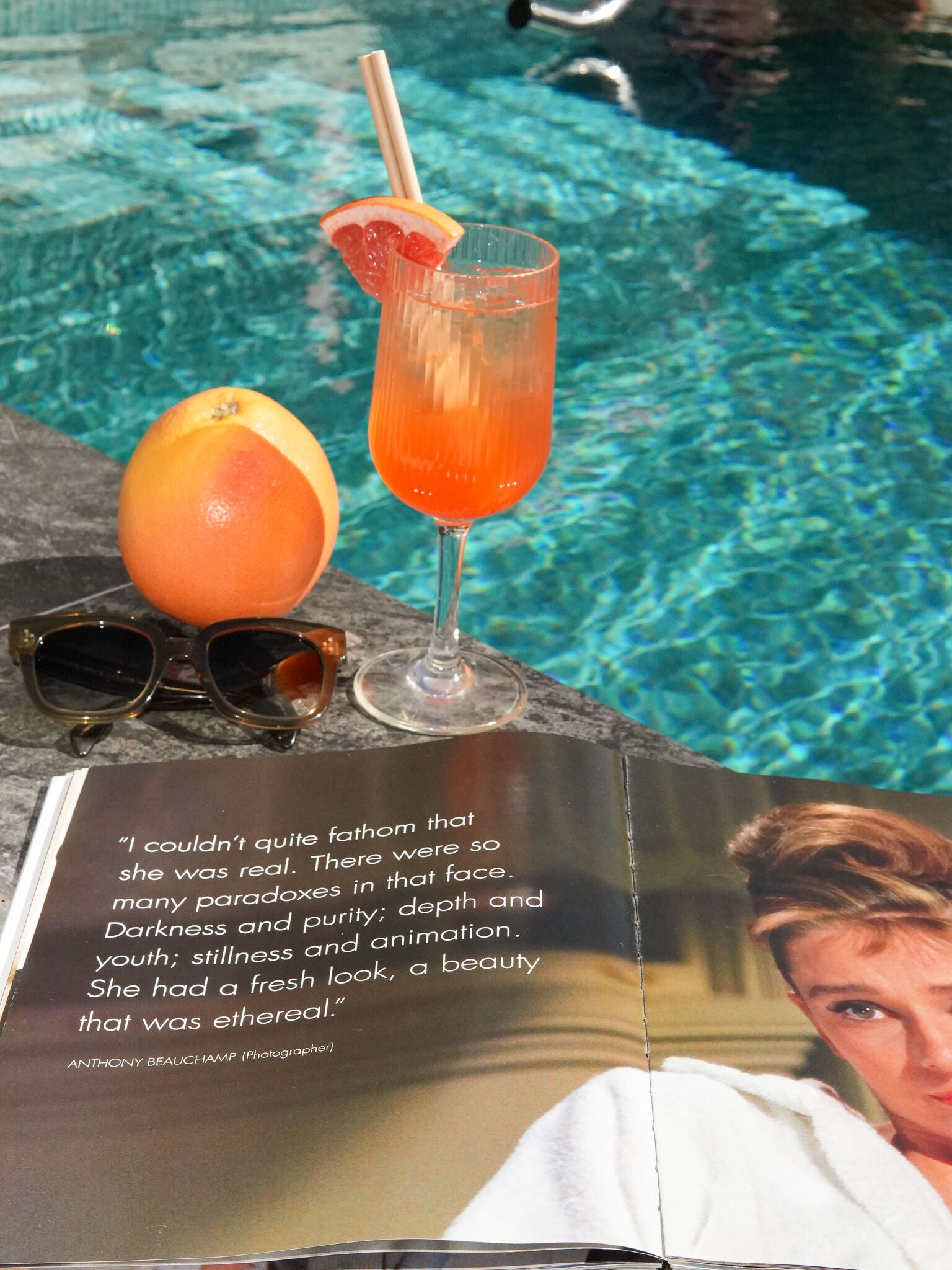 Orange cocktail i ett räfflat vinglas med grapefruktskiva vid en pool, bredvid solglasögon, en grapefrukt och ett öppet magasin.